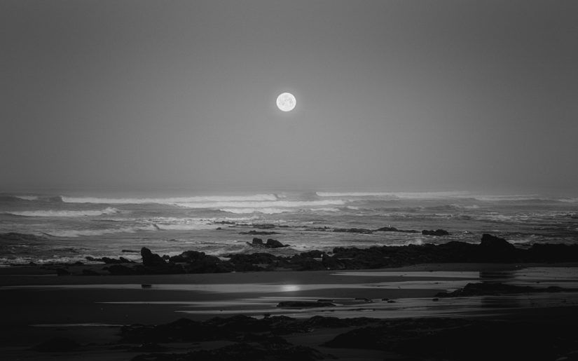 Black and white photo of moon on the horizon over an ocean beach.
